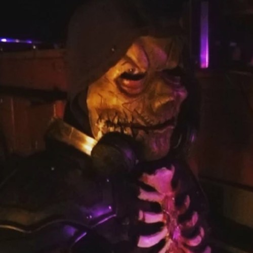 A person in a creepy skeleton mask and dark hood, wearing armor, poses in low light with purple hues in the background.