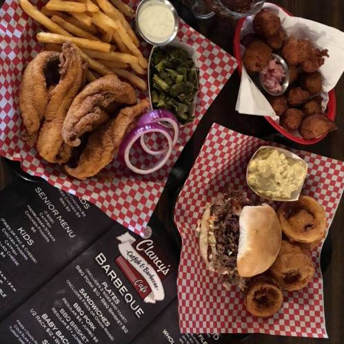 A spread of fried catfish, fries, collard greens, a pulled pork sandwich, onion rings, and hushpuppies on checkered trays.