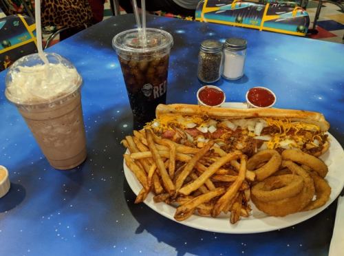 A plate of fries, onion rings, and a loaded hot dog, with a chocolate shake and a soda on a colorful table.