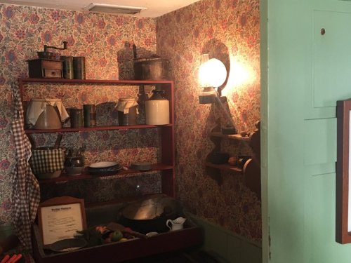 A vintage kitchen with floral wallpaper, shelves of pots, and a warm light fixture illuminating the space.