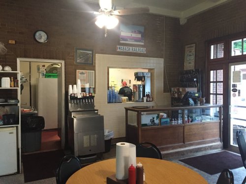 Interior of a cozy diner with a wooden counter, soda fountain, and a view of the kitchen. Wabash Railroad flag on the wall.