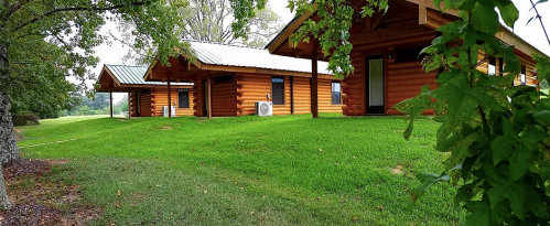 Three log cabins with metal roofs sit on a grassy area, surrounded by trees and greenery.