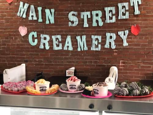 A display of various desserts, including ice cream cups and chocolate-covered treats, against a brick wall with a sign.