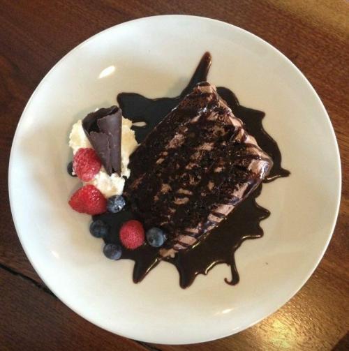 A slice of chocolate cake drizzled with sauce, served with whipped cream, raspberries, and blueberries on a white plate.