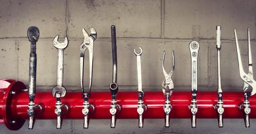 A row of various metal tools hanging on a red pipe against a gray wall.