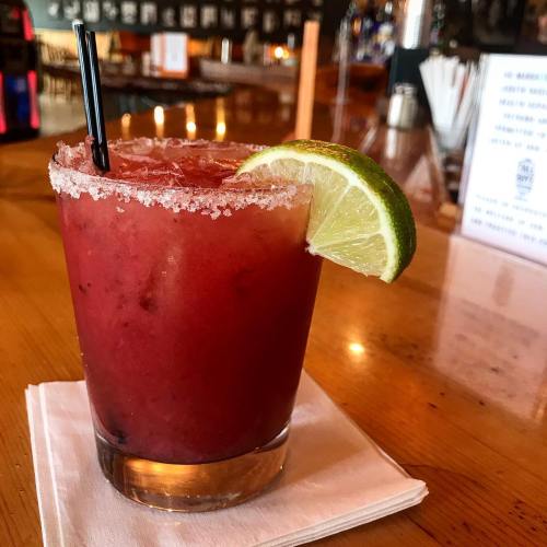 A vibrant red cocktail with a salted rim, garnished with a lime wedge, served on a wooden table.