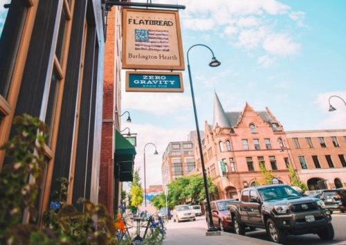 A street view featuring a restaurant sign, colorful flowers, and historic buildings under a blue sky with clouds.