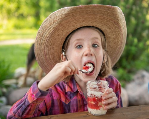 A child in a straw hat enjoys a dessert from a jar, with strawberries and cream, outdoors in a sunny setting.