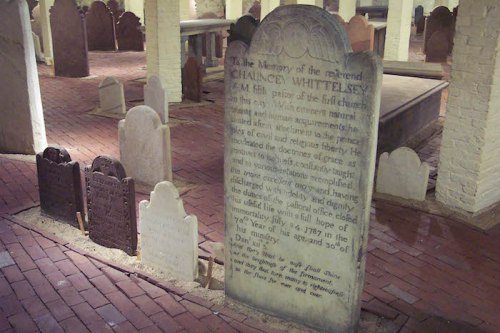 A historic grave marker in a dimly lit crypt, surrounded by smaller gravestones on a brick floor.