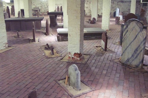 A dimly lit underground space with various old gravestones arranged on a brick floor.