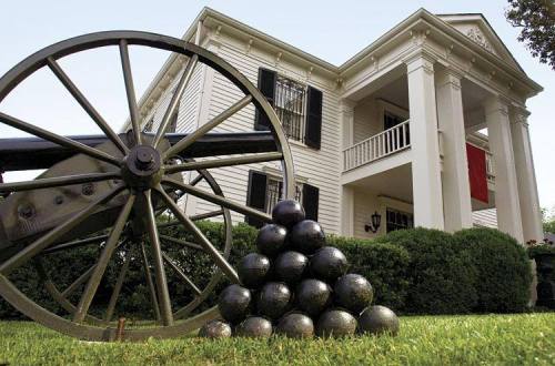 A historic cannon and cannonballs in front of a white, columned house with green bushes and a red flag.