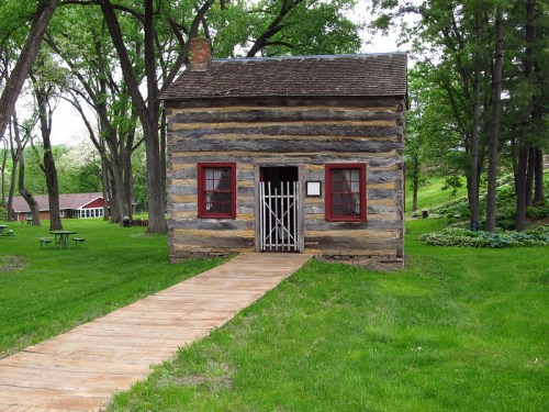 A small log cabin with red windows and a white gate, surrounded by green grass and trees, with a wooden path leading to it.