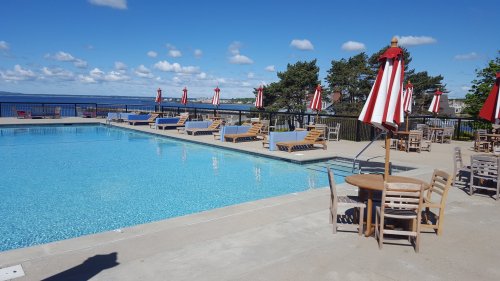 A sunny pool area with lounge chairs, red and white umbrellas, and a view of the water and blue sky.