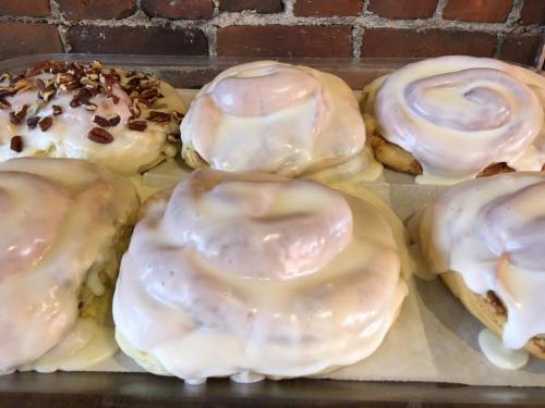 A tray of freshly baked cinnamon rolls topped with creamy icing and chopped nuts, set against a brick background.