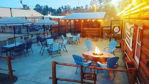 Outdoor seating area with blue chairs, tables, and a fire pit, surrounded by string lights and wooden structures.