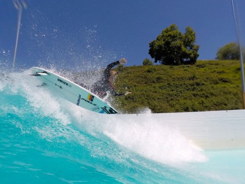 A surfer rides a wave in a pool, splashing water against a green hillside under a clear blue sky.