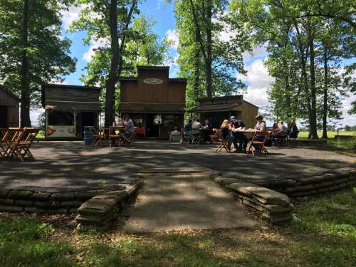 A rustic outdoor gathering space with people seated at tables, surrounded by trees and small buildings under a blue sky.