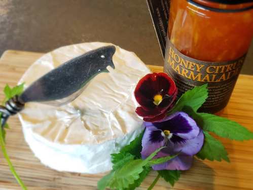 A round cheese wheel with a knife, honey citrus marmalade, and colorful edible flowers on a wooden board.