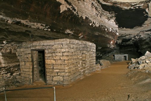 Interior of a cave featuring stone structures and sandy ground, with rugged rock formations above.