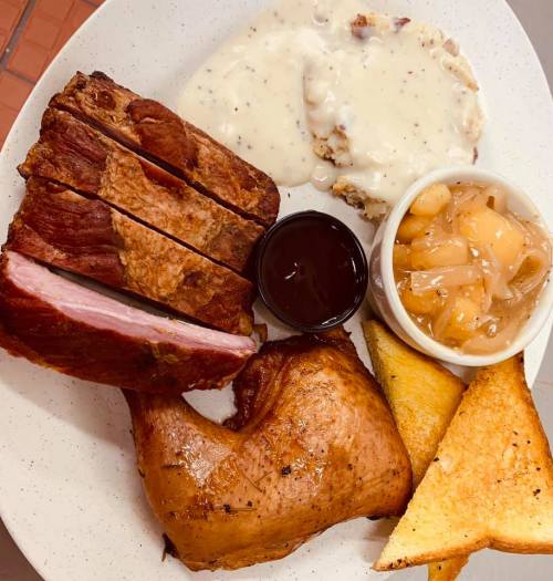 A plate featuring sliced smoked meat, a chicken leg, toast, baked beans, and creamy gravy over biscuits.
