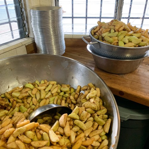 Two large bowls of sliced apples, one in a metal bowl with a serving spoon, and another in a colander on a wooden surface.