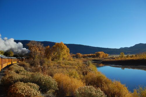 A scenic view of a train along a river, surrounded by autumn foliage and mountains under a clear blue sky.
