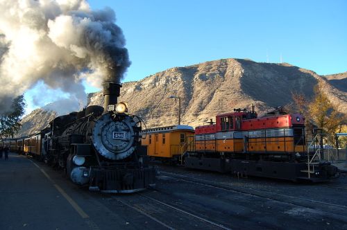 A steam locomotive emits smoke beside a diesel engine, with mountains in the background and a clear blue sky.