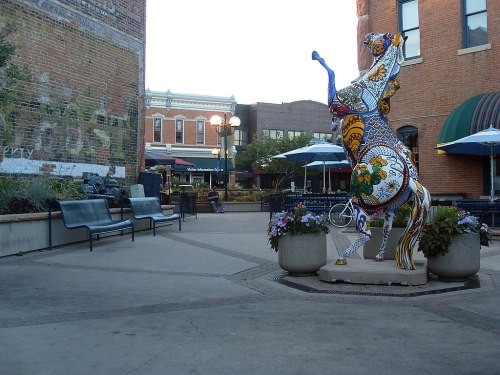 Colorful horse sculpture surrounded by flower pots in a lively outdoor plaza with benches and buildings in the background.