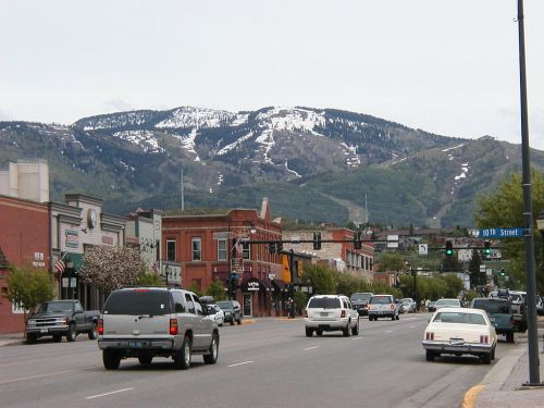A street scene with shops and cars, set against a backdrop of snow-capped mountains under a cloudy sky.