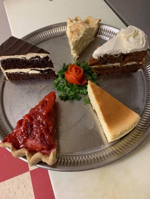 A silver platter with six slices of various cakes, including chocolate, carrot, cheesecake, and fruit pie, garnished with a rose.