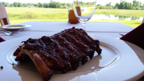 A plate of barbecue ribs sits on a table, with a scenic view of a field and water in the background.