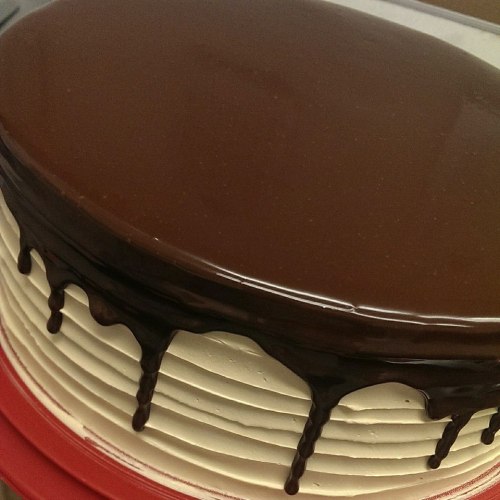 A round cake with a smooth chocolate ganache topping and decorative white frosting on the sides.