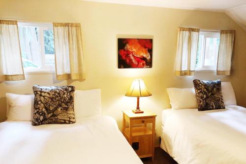 Cozy bedroom with two beds, decorative pillows, a lamp, and a colorful artwork on the wall. Natural light from windows.