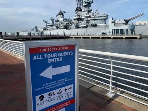 Sign directing tour guests to enter, with a battleship in the background and a waterfront view.