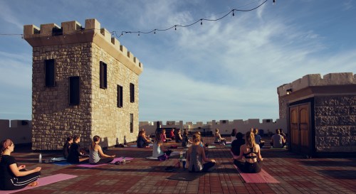 A group of people practicing yoga on a rooftop with castle-like architecture and string lights overhead.