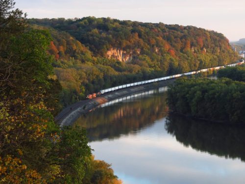 A scenic view of a river winding through lush green hills, with a train traveling along the bank at sunset.