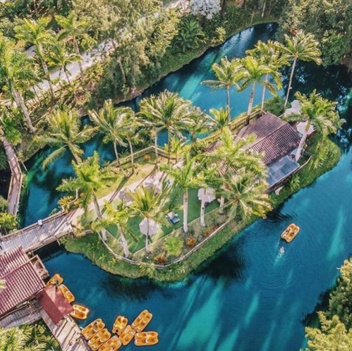 Aerial view of a tropical island with palm trees, a small building, and boats on a blue-green waterway.