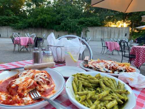 A table set outdoors with two plates of pasta, one with marinara and cheese, the other with pesto, and a drink.