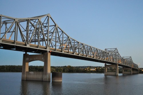 A steel truss bridge spans a river at dusk, with lights reflecting on the water below.