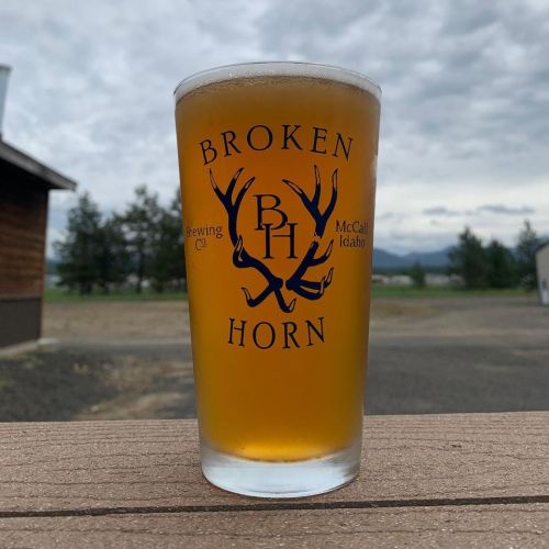 A pint glass of beer with the "Broken Horn Brewing Co" logo, set on a wooden table against a cloudy sky.