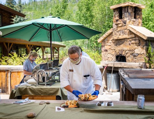 A chef in a white coat prepares a dish outdoors, while another person works in the background under an umbrella.