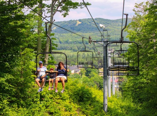 A family enjoys a scenic chairlift ride through lush green trees with mountains in the background.