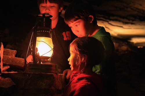Three children gathered around a lantern, illuminated by its warm glow in a dark, cave-like setting.