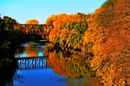 A serene river reflects vibrant autumn foliage and a bridge under a clear blue sky.