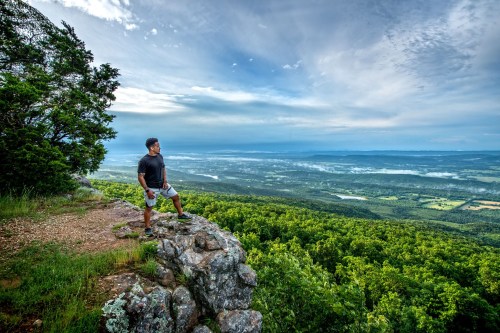 A person stands on a rocky outcrop, overlooking a lush green valley and distant mountains under a cloudy sky.