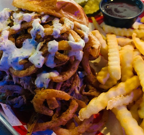 A plate featuring a pulled pork sandwich topped with onion rings and sauce, alongside crinkle-cut fries and pickles.