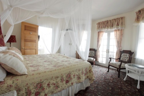 Cozy bedroom with a floral bedspread, sheer canopies, vintage chairs, and natural light from large windows.