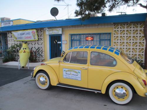 A yellow vintage car parked outside a colorful ice cream shop with a banana mascot and a "liquor" sign.