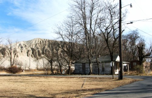 A barren landscape with a dirt hill in the background, a few bare trees, and an old wooden house near a road.