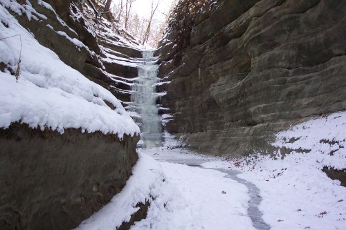 A frozen waterfall cascades down rocky cliffs, surrounded by snow-covered ground and trees in a winter landscape.